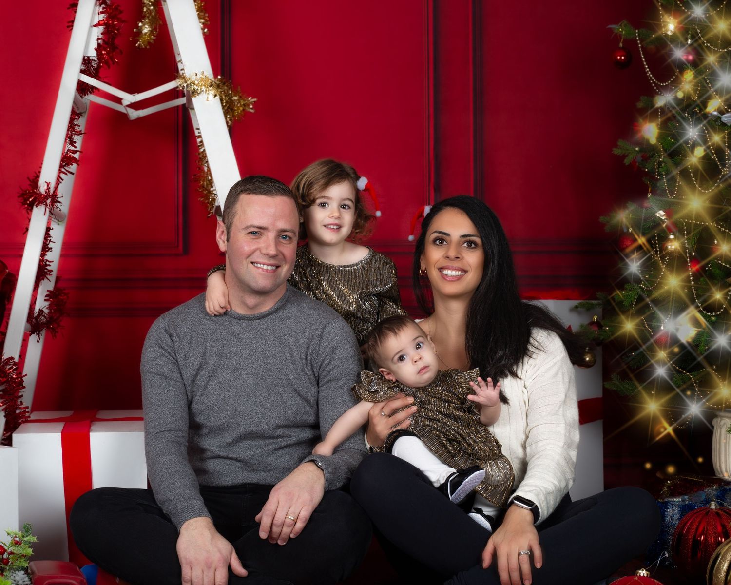 Family of four posing for a festive Christmas photo with a decorated tree and red holiday backdrop.