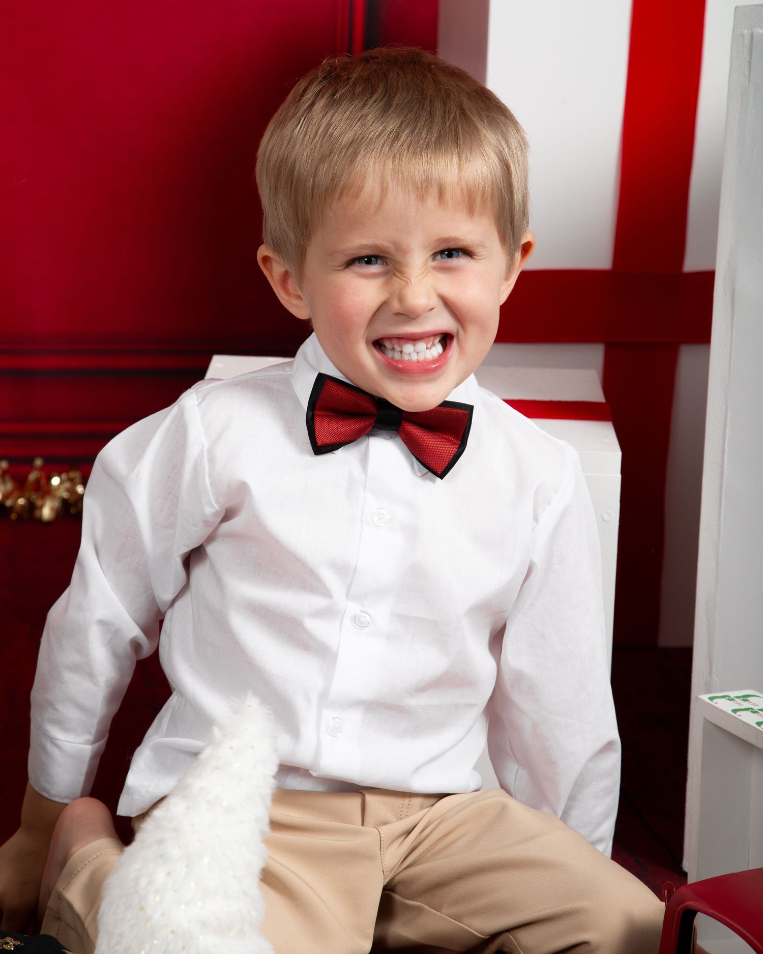 A cheerful young boy in a white dress shirt and red bow tie smiles broadly during a festive holiday photo session.