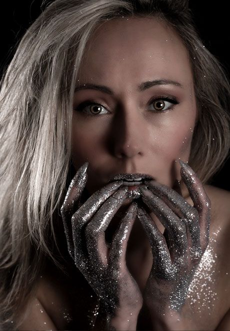 Blonde woman with glitter-covered hands held to lips in dramatic dark studio beauty portrait.
