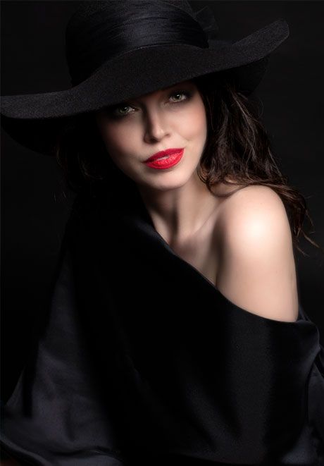 Glamorous woman in wide-brim black hat and off-shoulder black wrap with red lips on dark background.