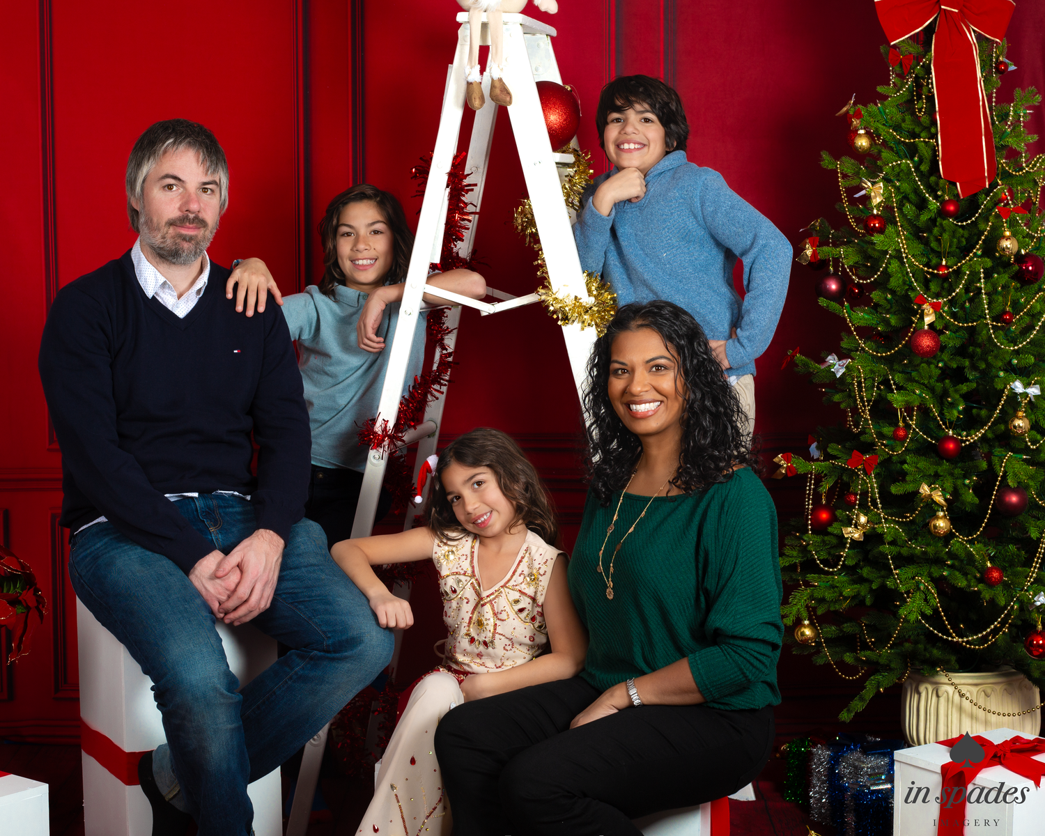 Family of five posing for a Christmas portrait with a decorated tree and festive red background.