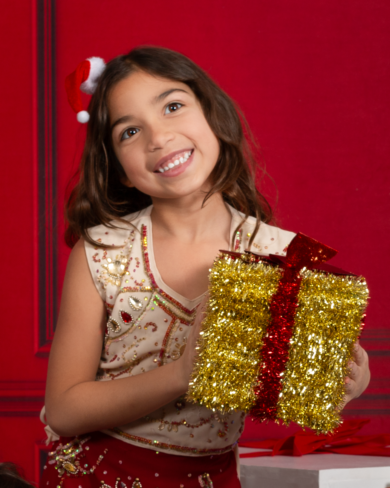 Smiling young girl in festive outfit holding a gold and red tinsel gift box against a red Christmas background.