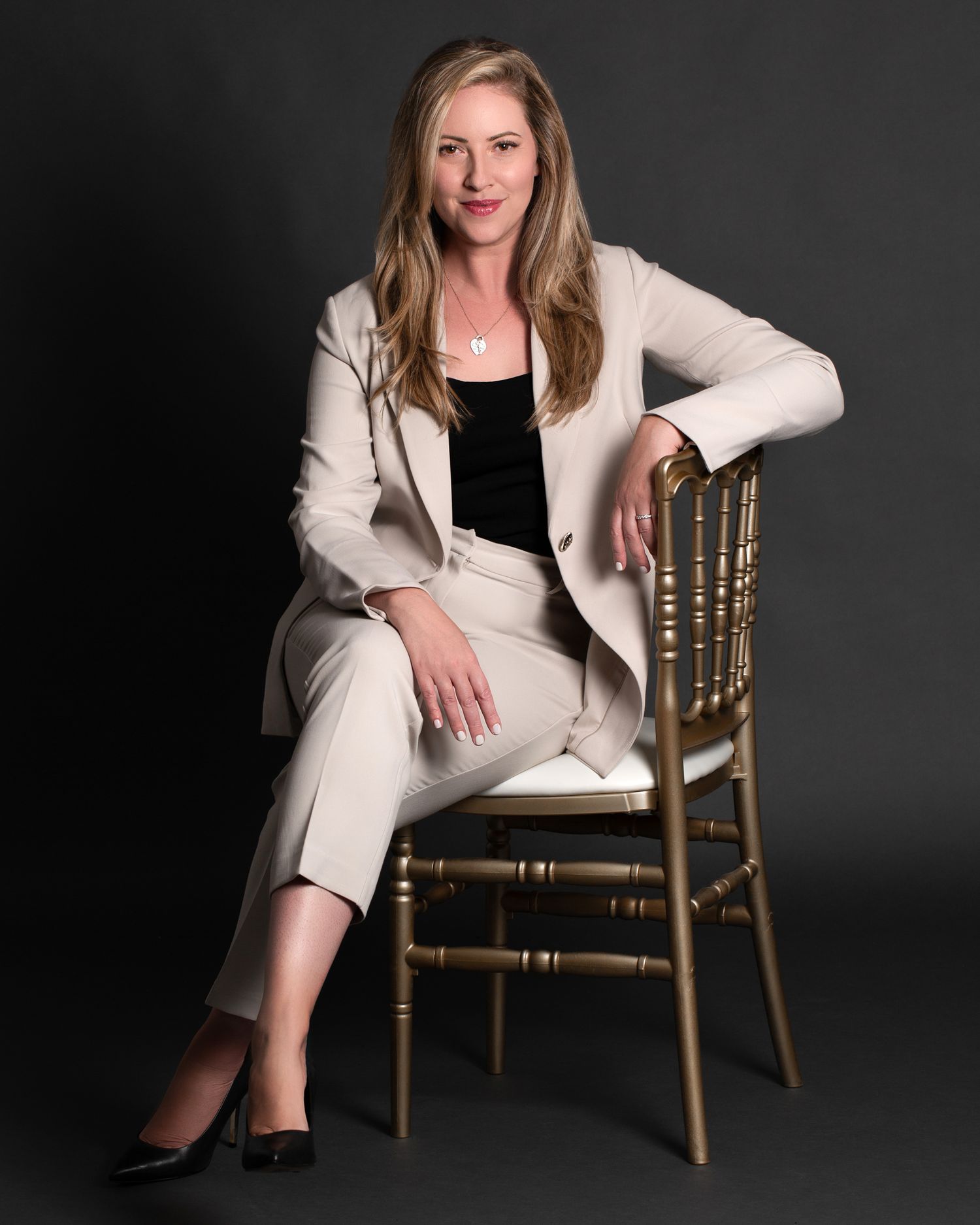 Professional woman in beige suit seated on gold chair against dark gray studio background. Ottawa real estate branding headshot photography