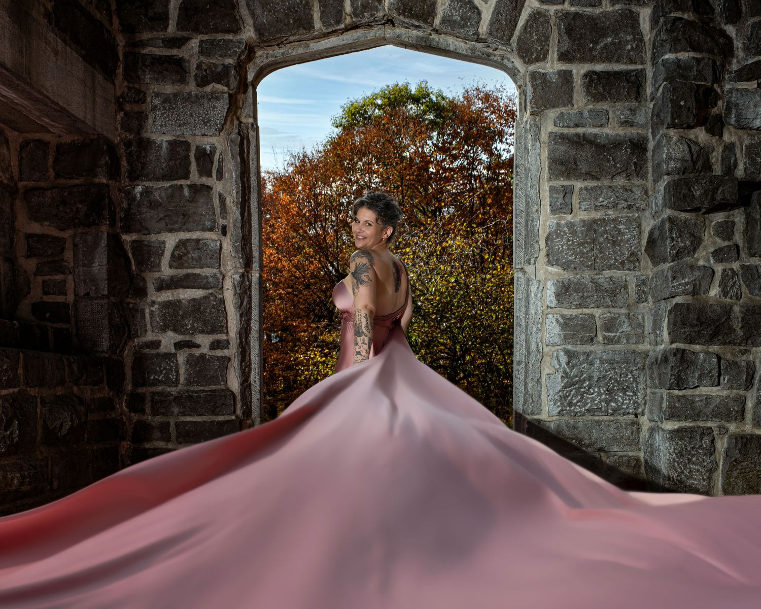 Woman in pink flowing gown stands in stone archway with autumn trees.  Ottawa flying dress photography. Mackenzie King estate