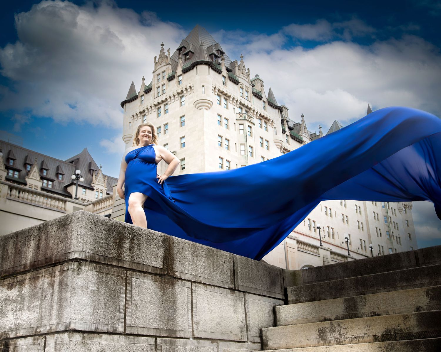 Woman in flowing royal blue gown poses on stone steps with chateau laurier  building backdrop. Ottawa flying dress photography.