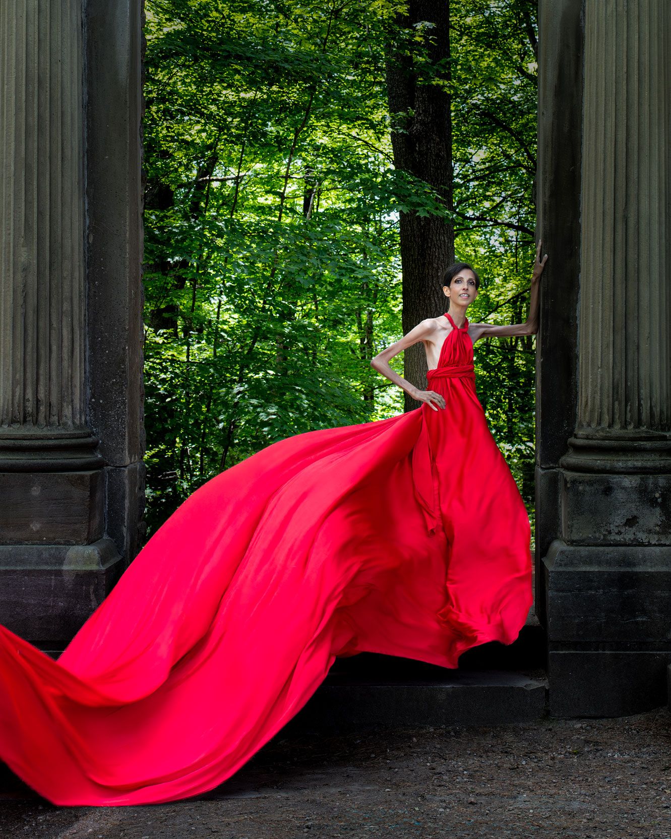 Elegant woman in dramatic red flying dress poses between stone columns in a lush green forest. Ottawa flying dress photography.