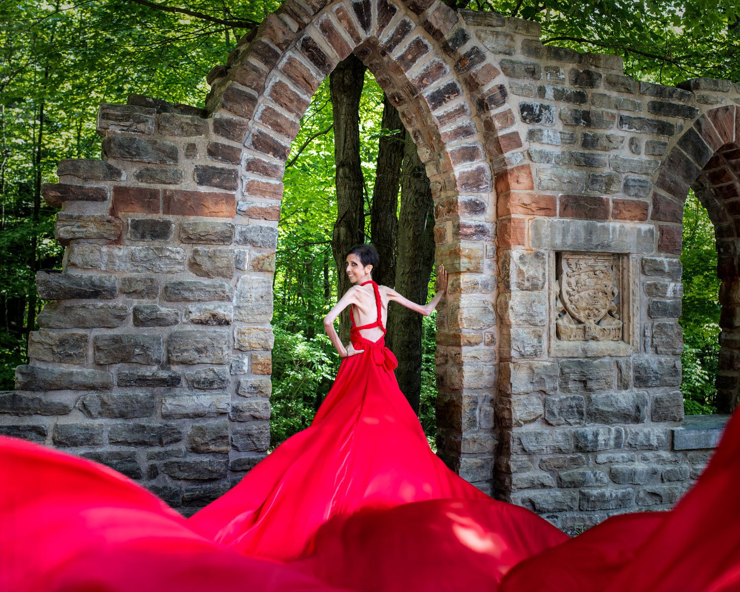 Woman in red flying dress poses before ornate stone arch in forest.  Ottawa flying dress photography. Mackenzie King estate