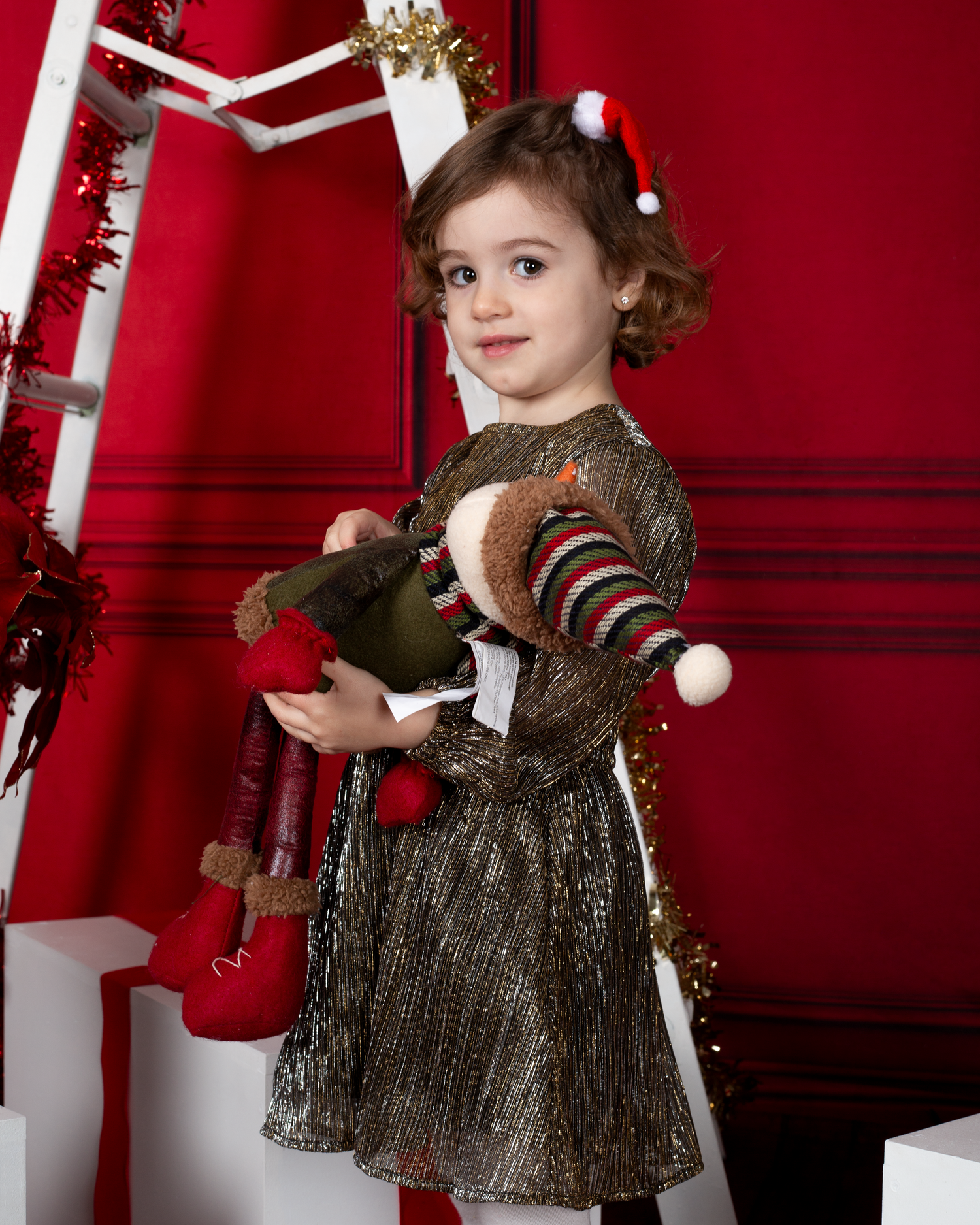 A toddler girl in a gold metallic dress holds a stuffed reindeer toy in a festive red Christmas-themed studio setting.