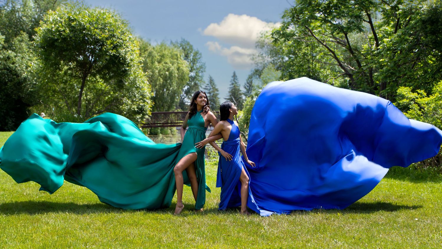 Two women in dramatic blue and green flying dresses laugh together in sunny experimental farm. Ottawa flying dress photography.