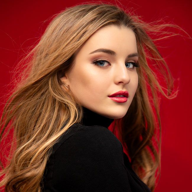 Beautiful young woman with flowing auburn hair and red lips in black turtleneck against red background.