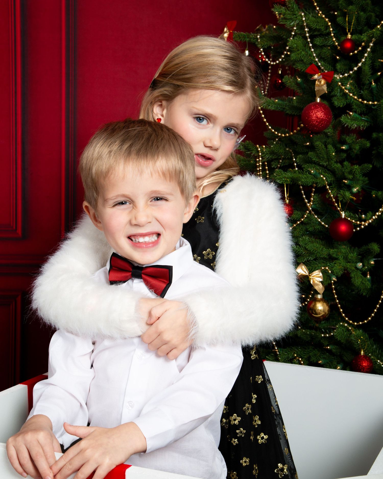 Two festive children in holiday attire pose together in front of a decorated Christmas tree with red ornaments.