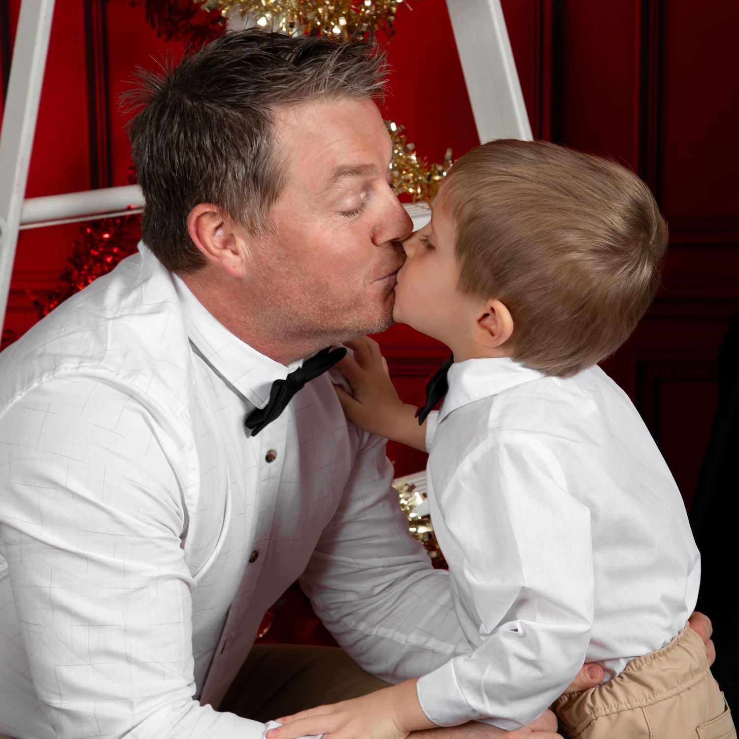 A man in a white dress shirt and bow tie shares a sweet kiss with a young boy during a festive Christmas photo session.