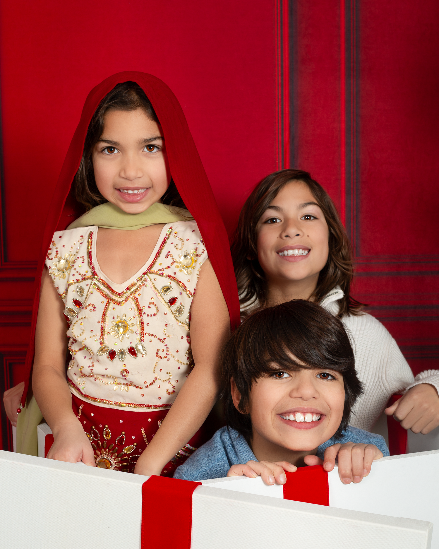 Three smiling children pose in front of a red background with a white gift box adorned with a red ribbon.