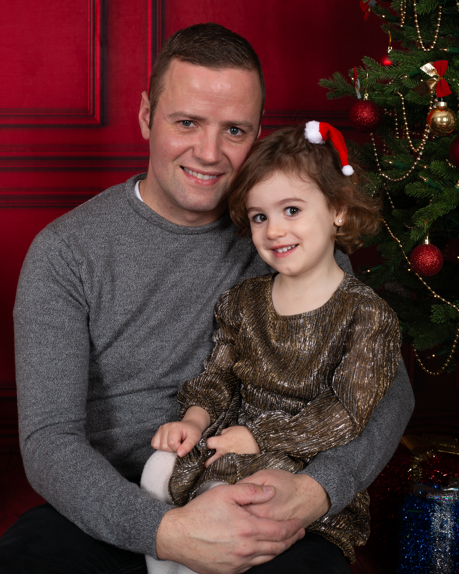 A man and young girl pose together in front of a decorated Christmas tree, smiling for a festive holiday portrait.