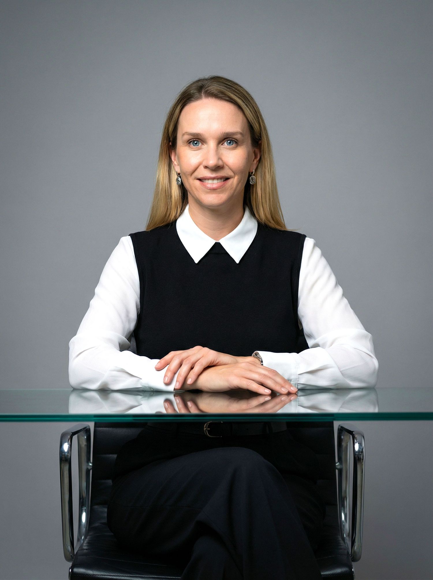 A professional blonde woman in a black vest and white shirt sits at a glass desk against a gray background.