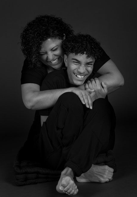 Black and white portrait of laughing mother hugging teenage son on studio floor.