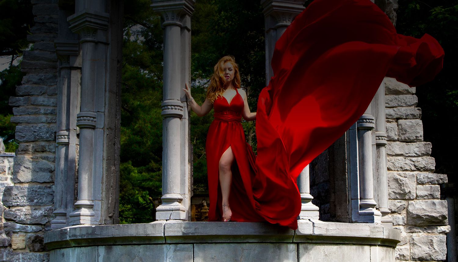 Woman in flowing red gown standing among stone columns with dramatic fabric movement outdoors.
