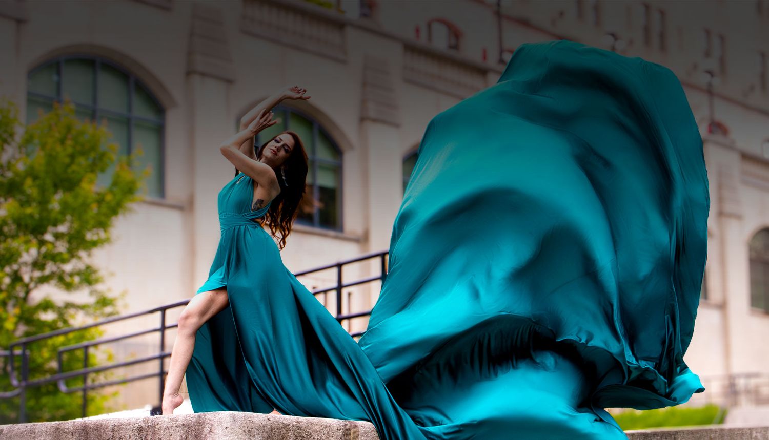 Woman in teal flowing dress with dramatic fabric movement seated on outdoor stone steps.