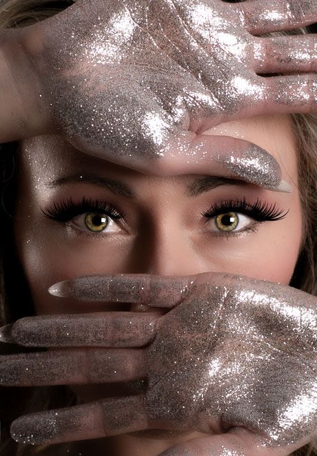 Close-up of woman with striking green eyes framed by glitter-covered hands.