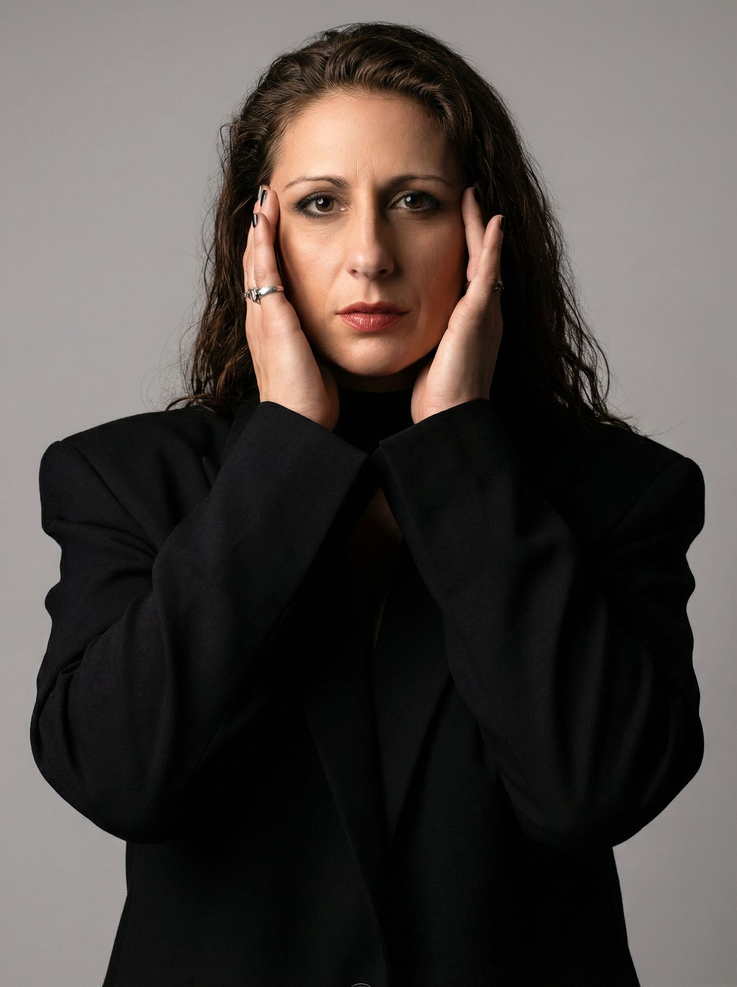 Woman in black jacket pressing hands to temples, stressed expression, gray background.