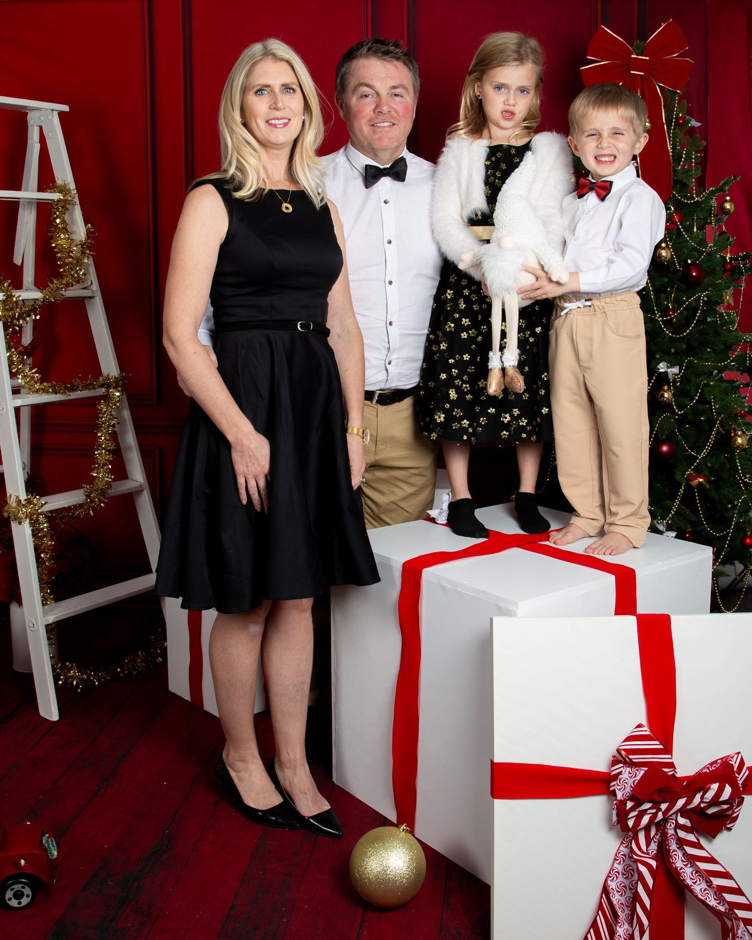 Family of four posing for a festive Christmas photo with gifts and decorated tree on red background.