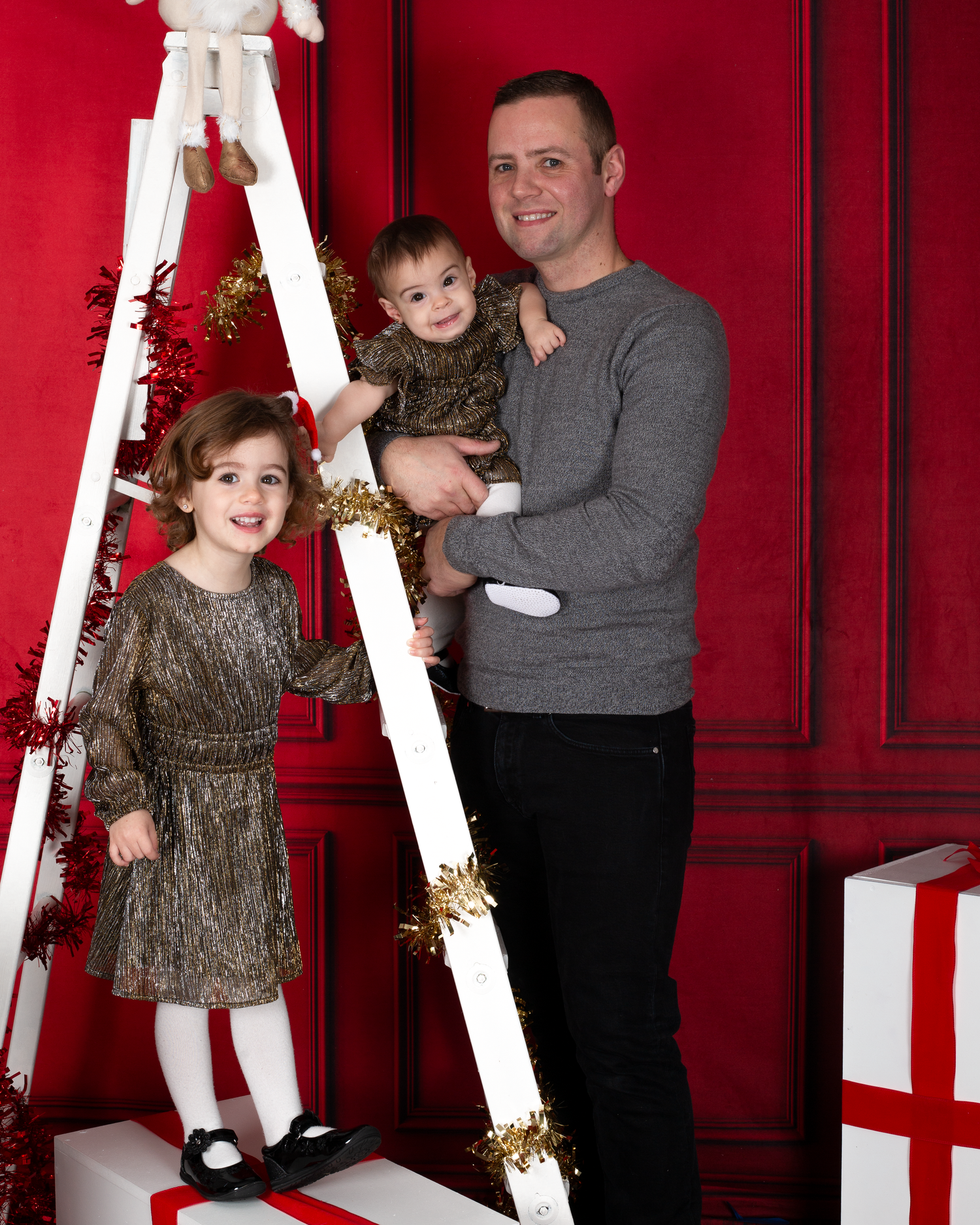 A man holds a baby while a young girl poses beside a decorated white ladder in a festive red Christmas photo studio.