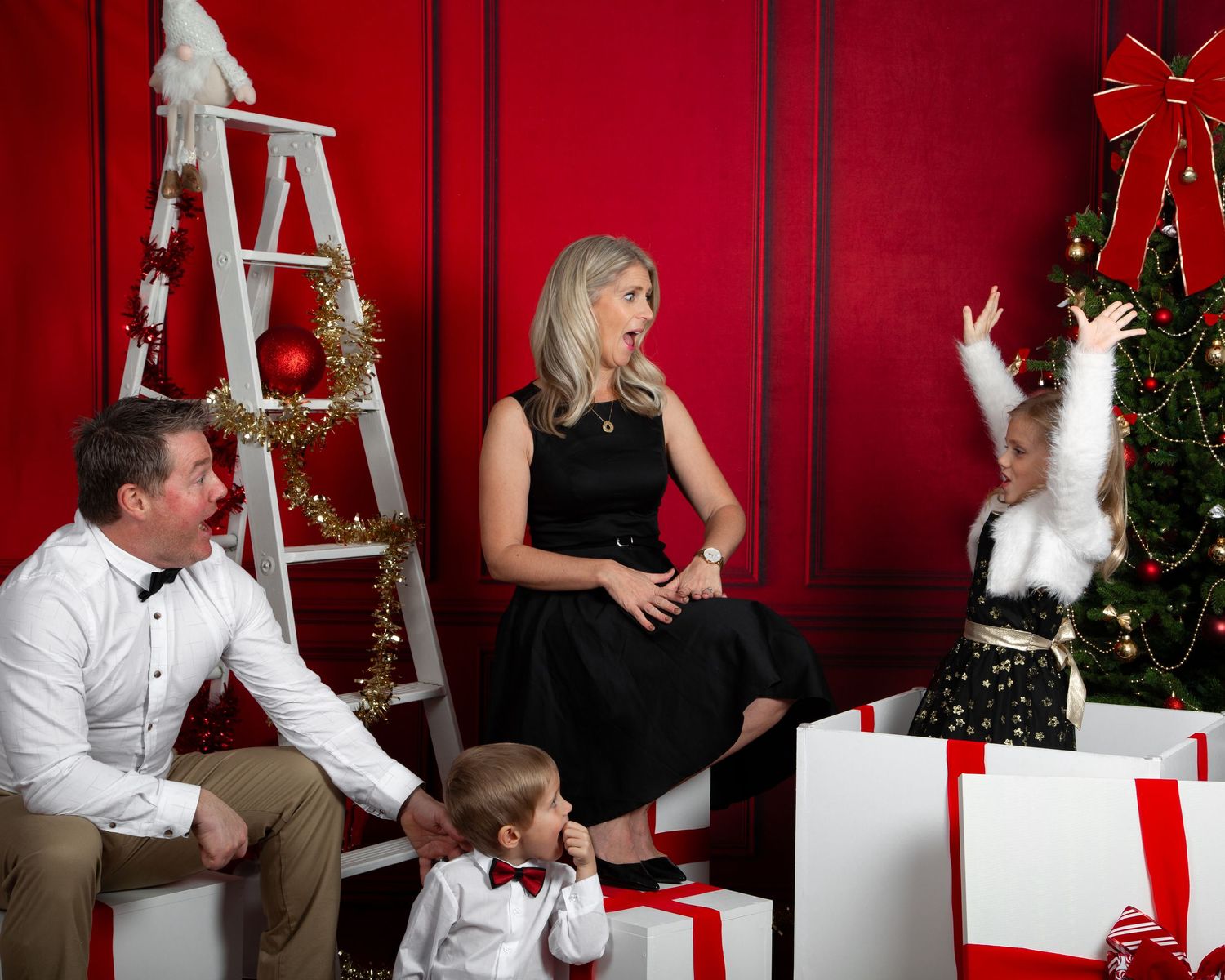 Family Christmas photo with parents and two children posing near decorated tree and white gift boxes on red background.