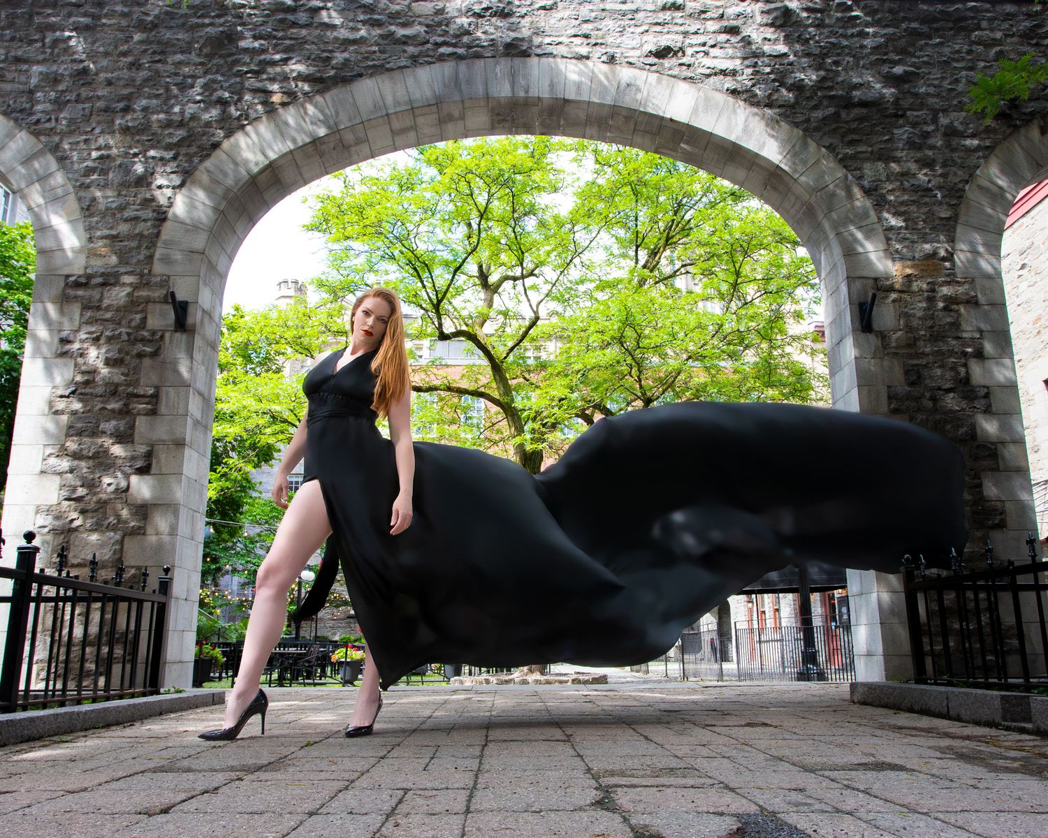 Redhead woman in black flying dress strikes a bold pose under a historic stone archway. Ottawa flying dress photography. Byword market