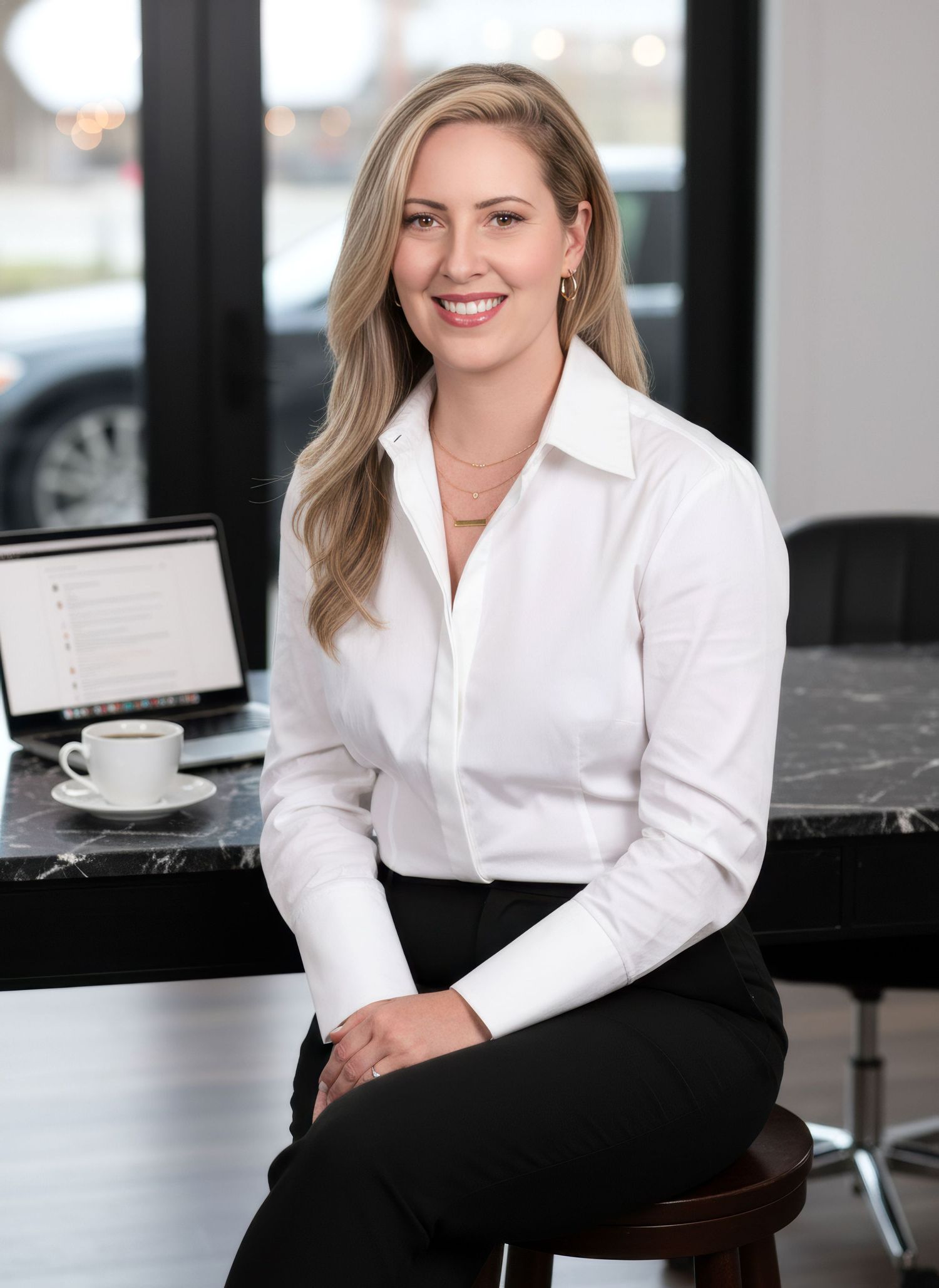 Smiling blonde woman in white shirt seated at marble table with laptop and coffee.