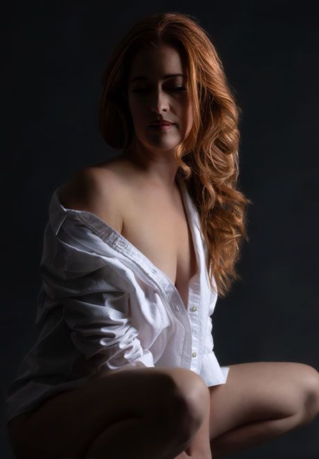 Redhead woman in open white button-down shirt seated in moody dark studio portrait photography.