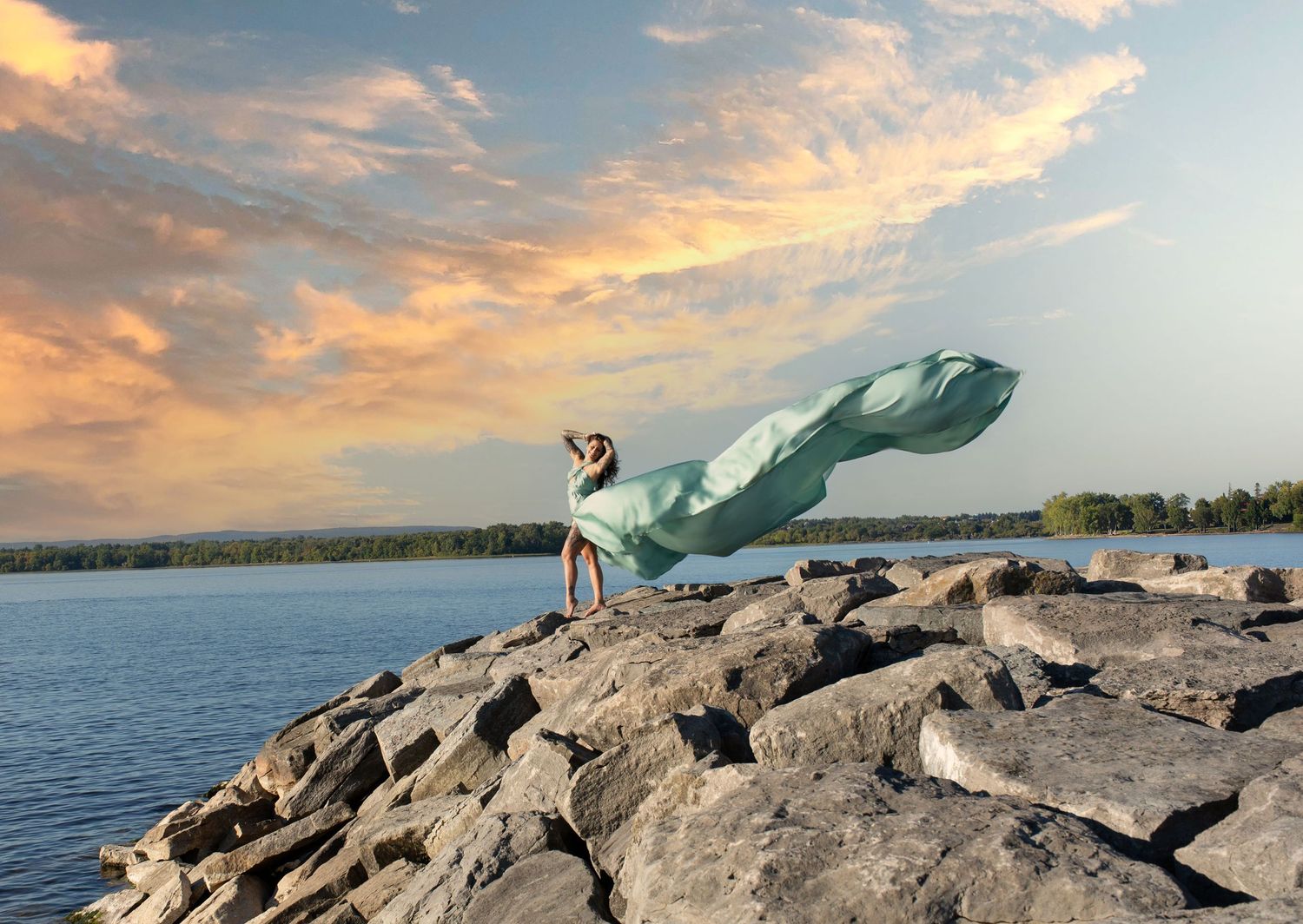 Woman in flowing safe dress on rocky shoreline with lake and dramatic sunset sky. Ottawa flying dress luxury photography. Britannia beach
