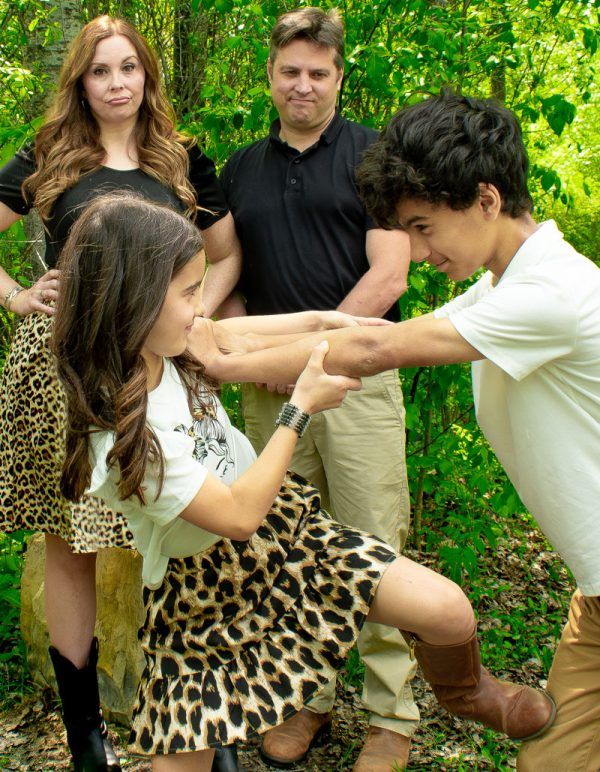 Family portrait in nature with parents watching two children playfully interact in a wooded outdoor setting.