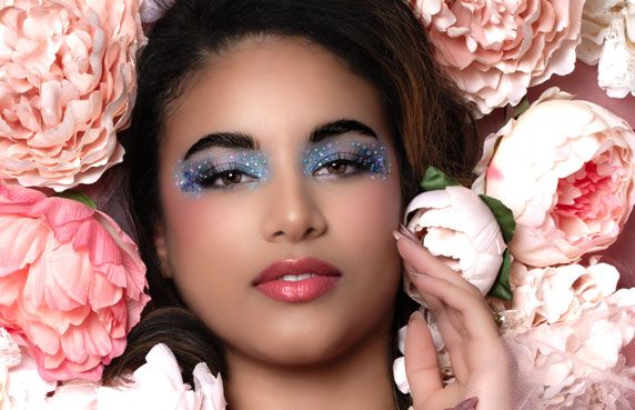 Young woman with blue glitter eyeshadow surrounded by pink peonies in a beauty editorial.