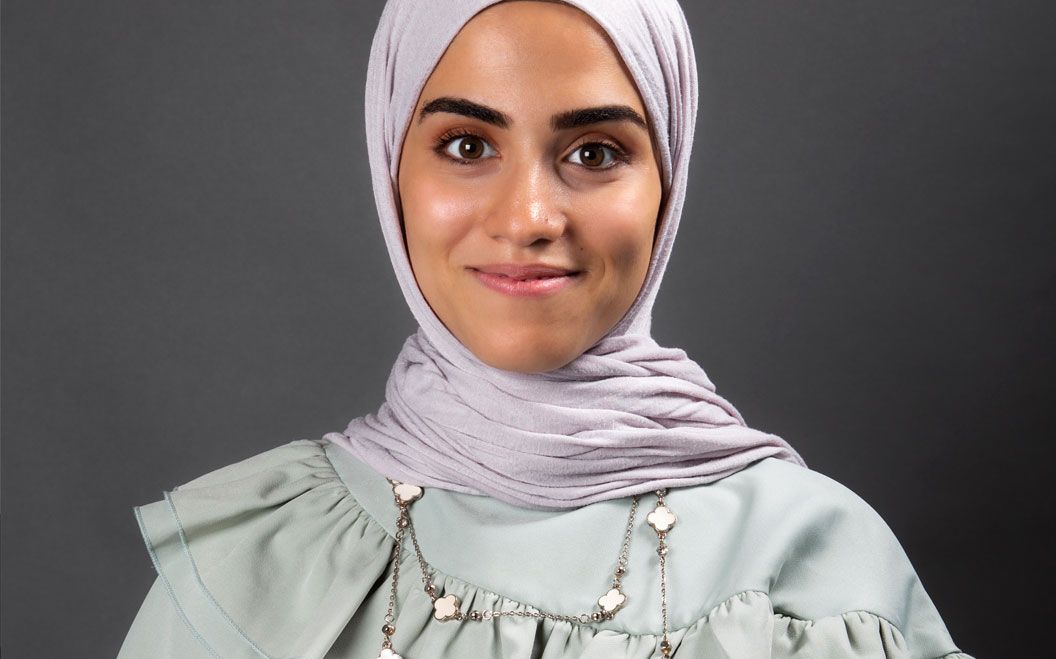 Young woman wearing lavender hijab and mint green top smiling against gray background.