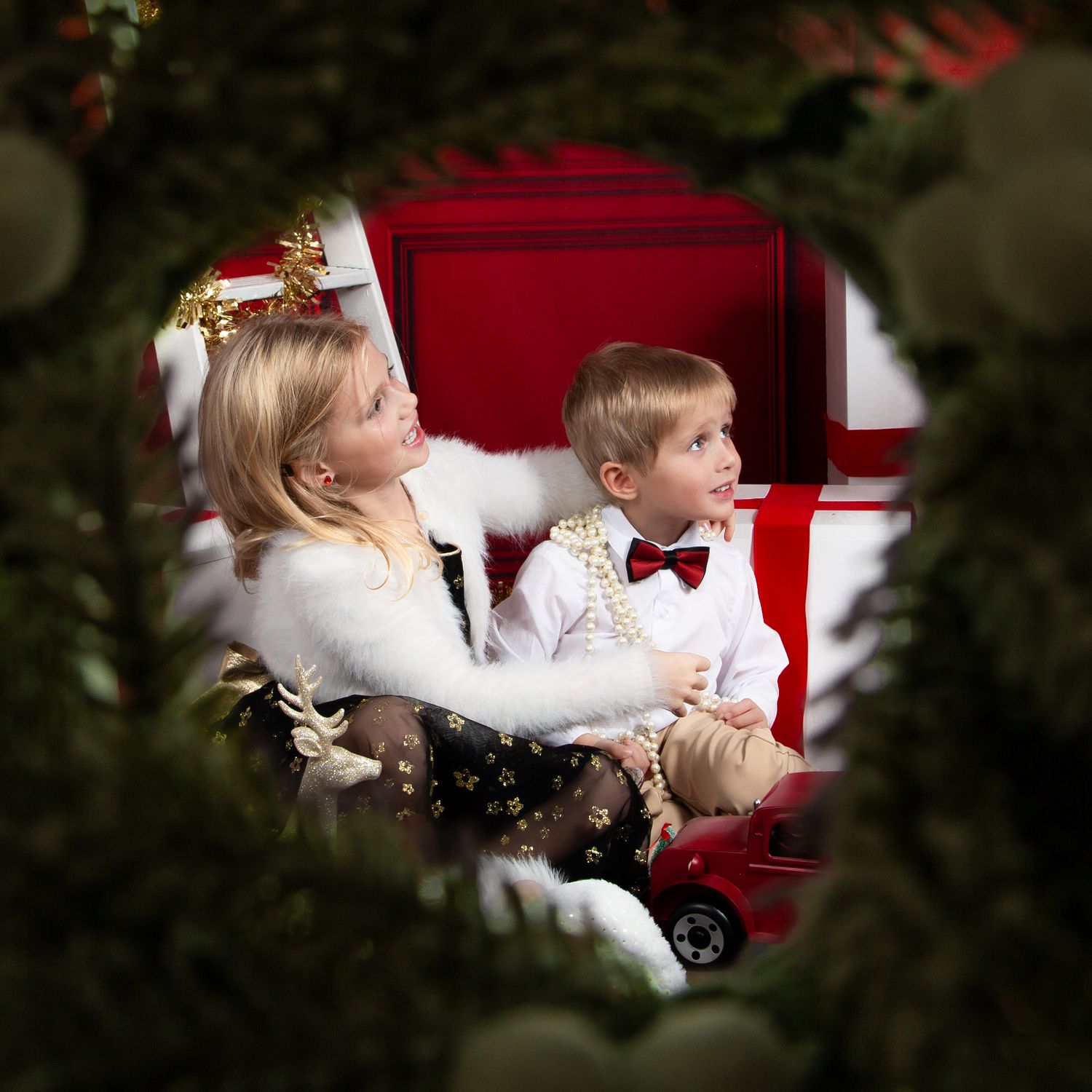 Two children in holiday attire sit together near a Christmas tree, with a red backdrop and festive decorations.