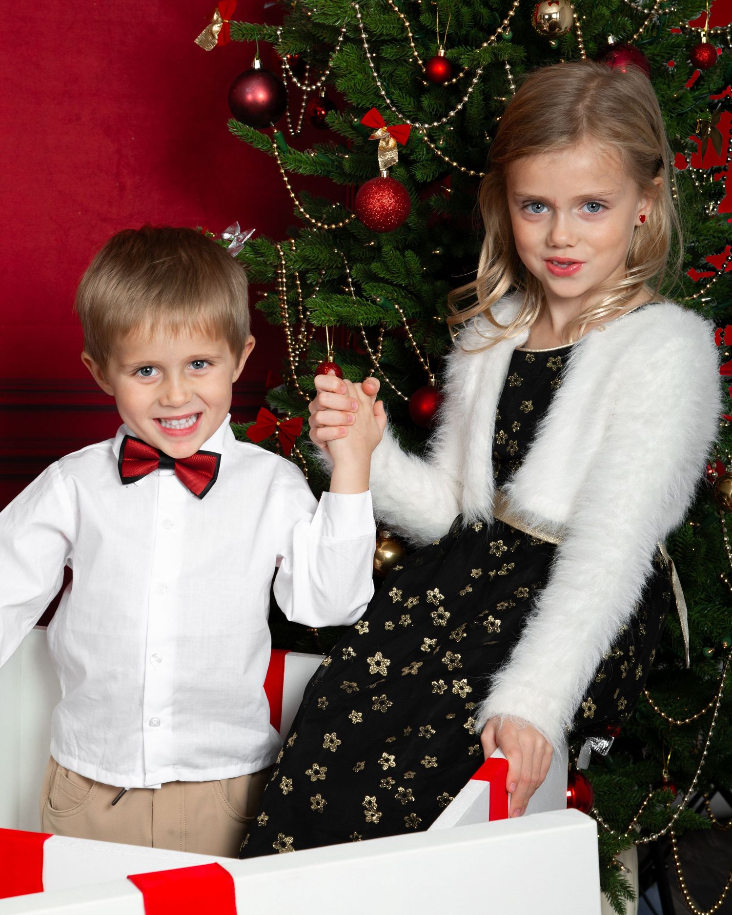 Two children in festive holiday outfits pose beside a decorated Christmas tree with gifts.