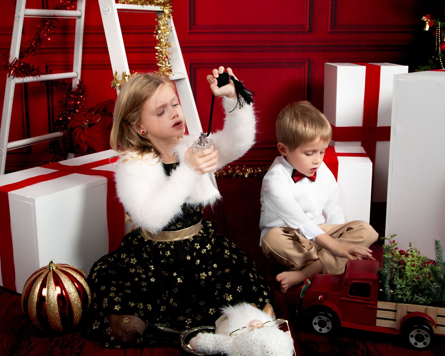 Two children in holiday attire pose with Christmas gifts and decorations against a festive red background.