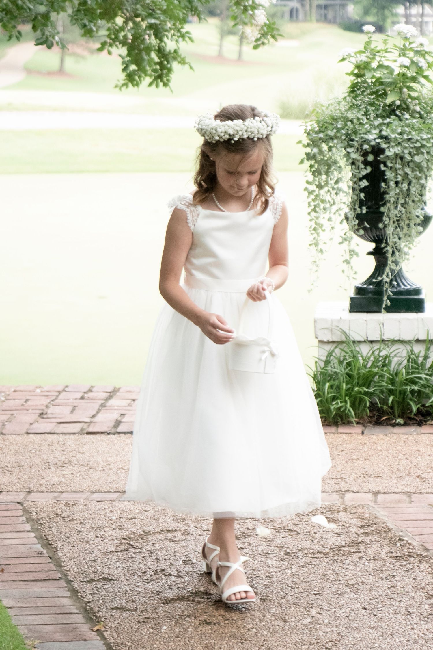 A flower girl in a white dress and floral crown walks down a garden path.