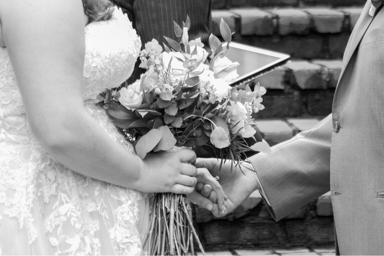 A black and white close-up of hands holding a wedding bouquet during the ceremony. - every detail