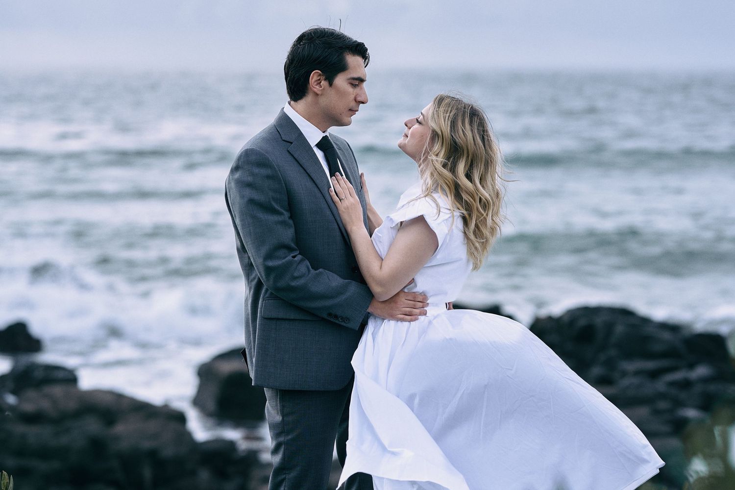 Romantic couple poses on rocky beach shoreline in wedding attire.