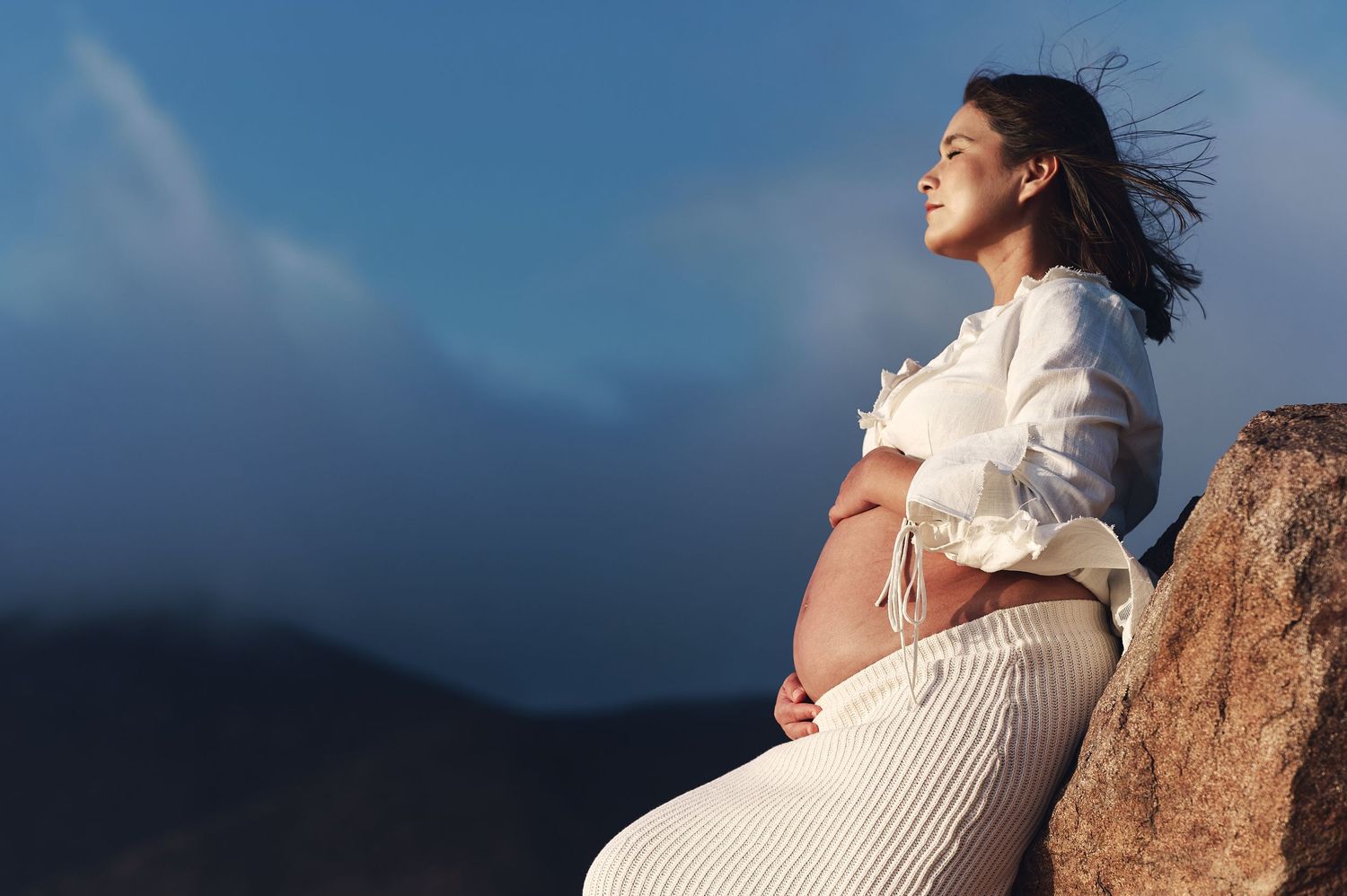 Maternity photo of an expectant mother in white clothing against a mountain backdrop.