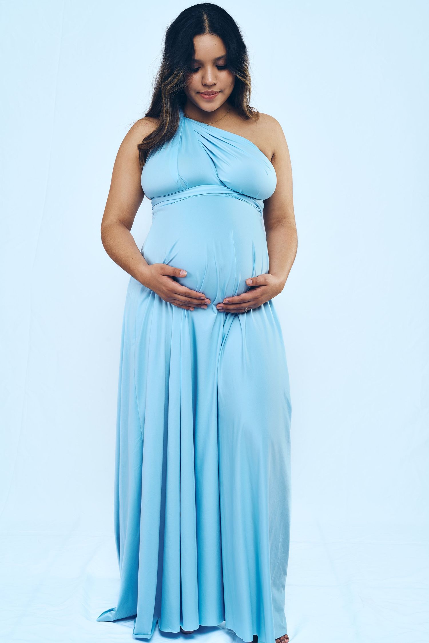 Woman in light blue flowing dress poses against white background showing pregnancy.