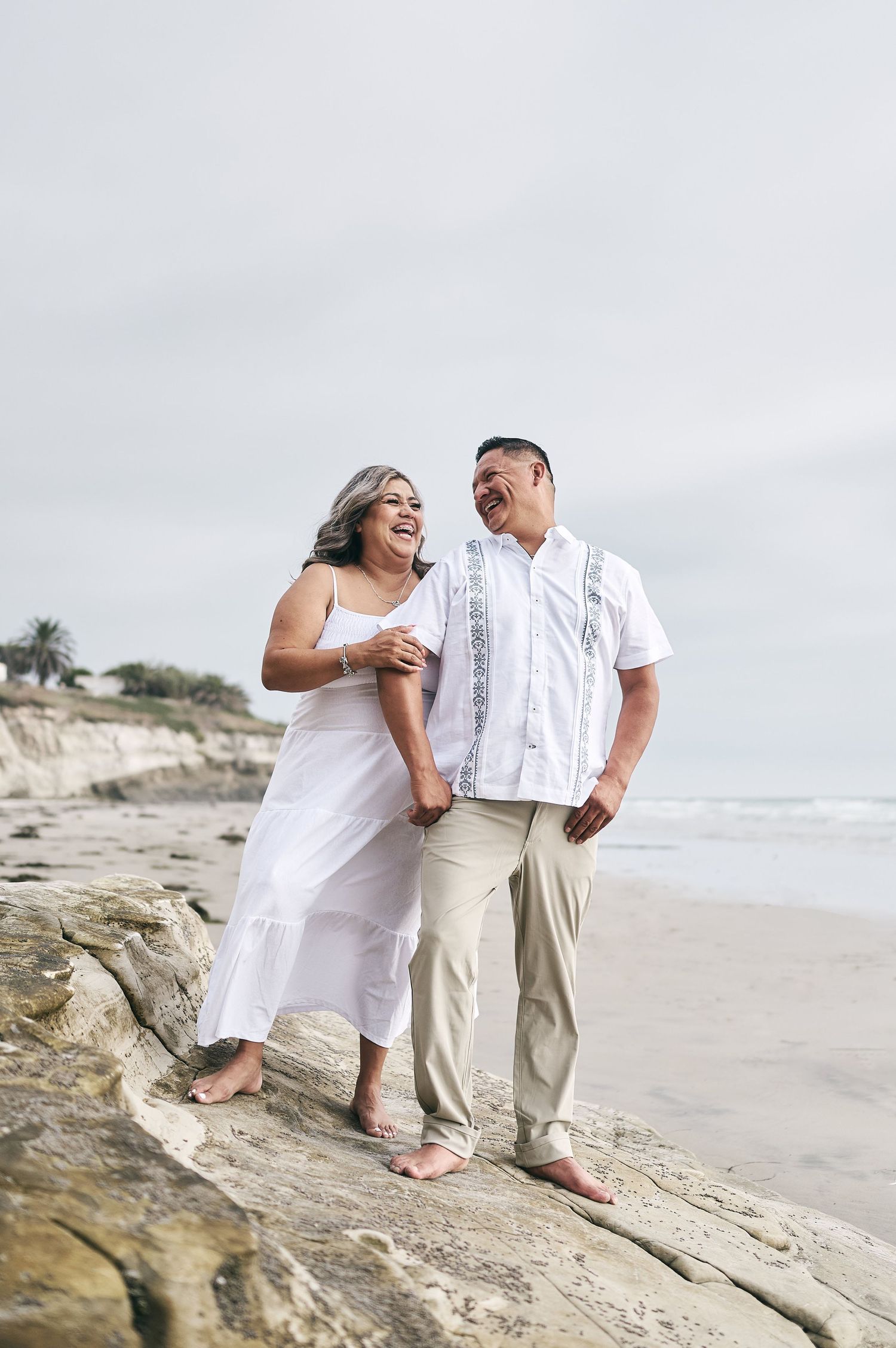 A couple shares joyful moments on rocky beach formations while wearing white and beige clothing.