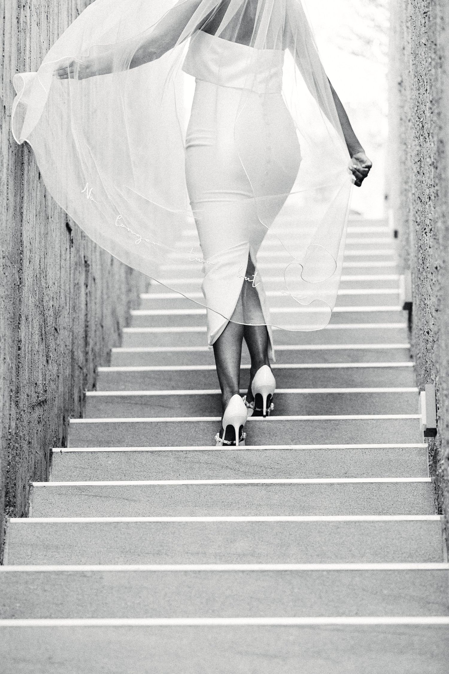 Black and white photo of bride ascending concrete stairs while holding flowing veil.