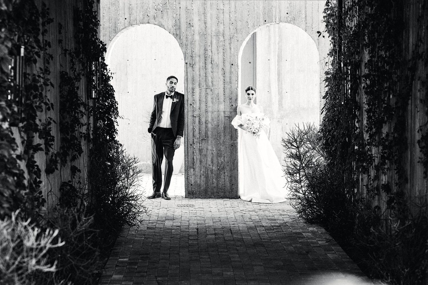 Black and white wedding photo of couple standing in separate arched doorways with ivy.