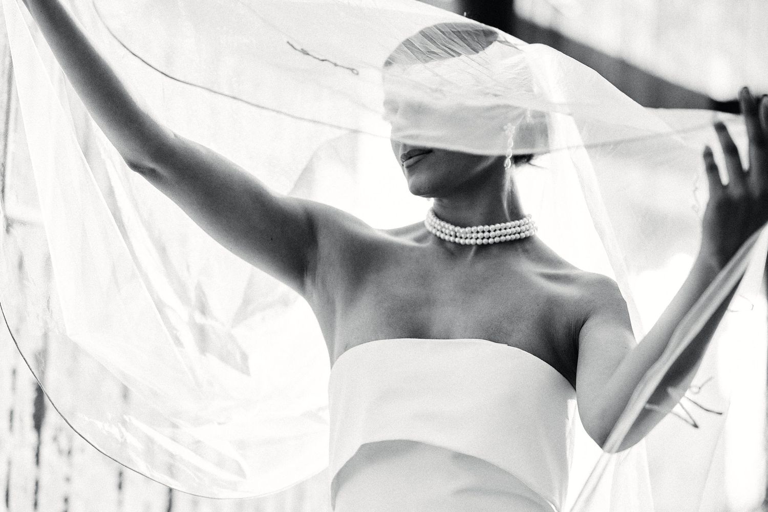 Artistic black and white photo of bride holding sheer veil against bright light.