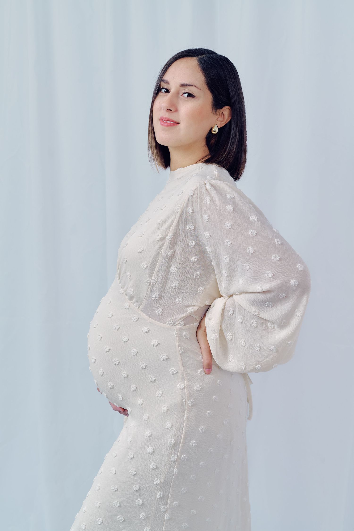 Studio maternity portrait in a white textured dress with balloon sleeves against light backdrop.