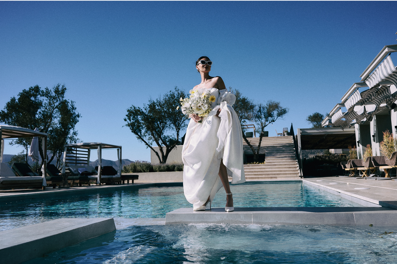 A bride in a flowing white dress sits elegantly by a serene swimming pool on a sunny day.