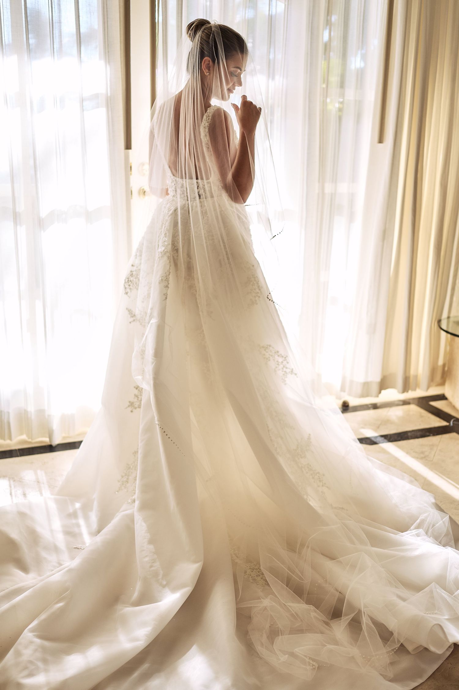 Elegant wedding dress with long train photographed in a sunlit room with sheer curtains.