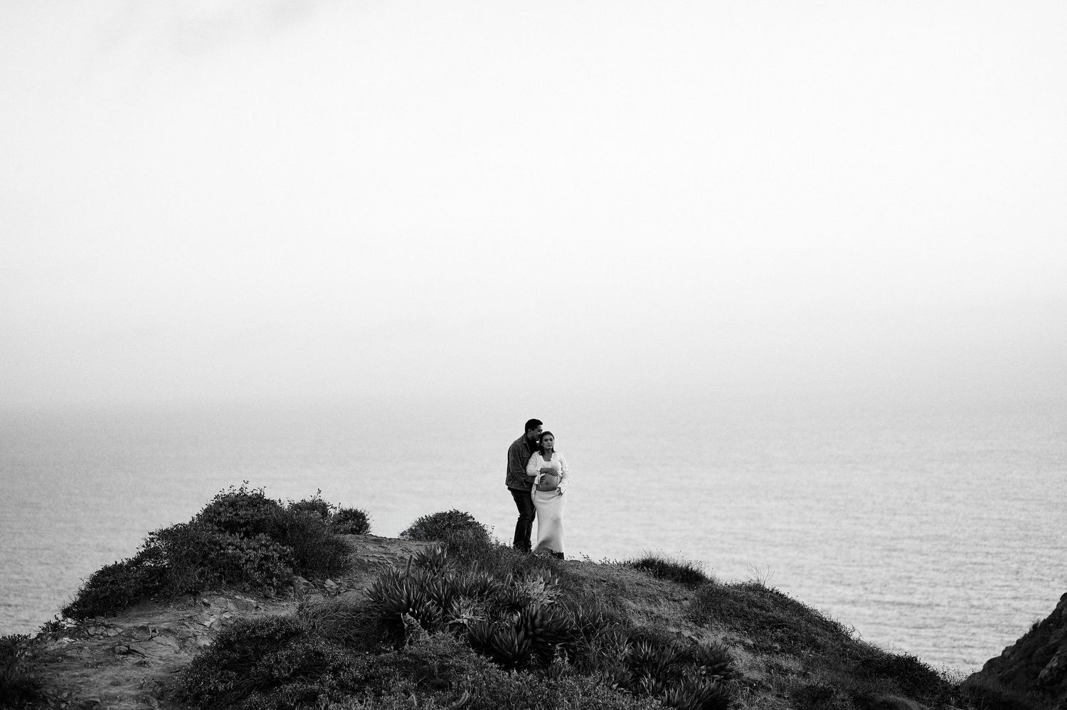 A silhouetted figure stands on a cliff edge overlooking a misty ocean horizon in black and white.